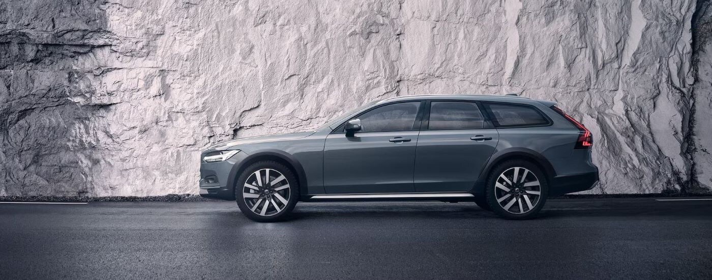 A silver 2023 Volvo V90 Cross Country is shown from the side in front of a rock wall.
