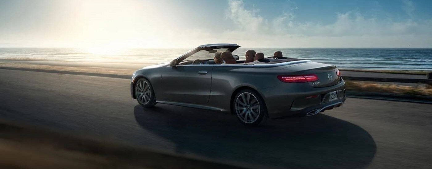 A grey 2022 Mercedes E-Class Cabriolet is shown from the side driving past the ocean.
